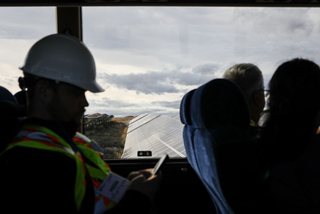 People sitting in seats in a bus pass by a grid of solar panels, in focus in the background.