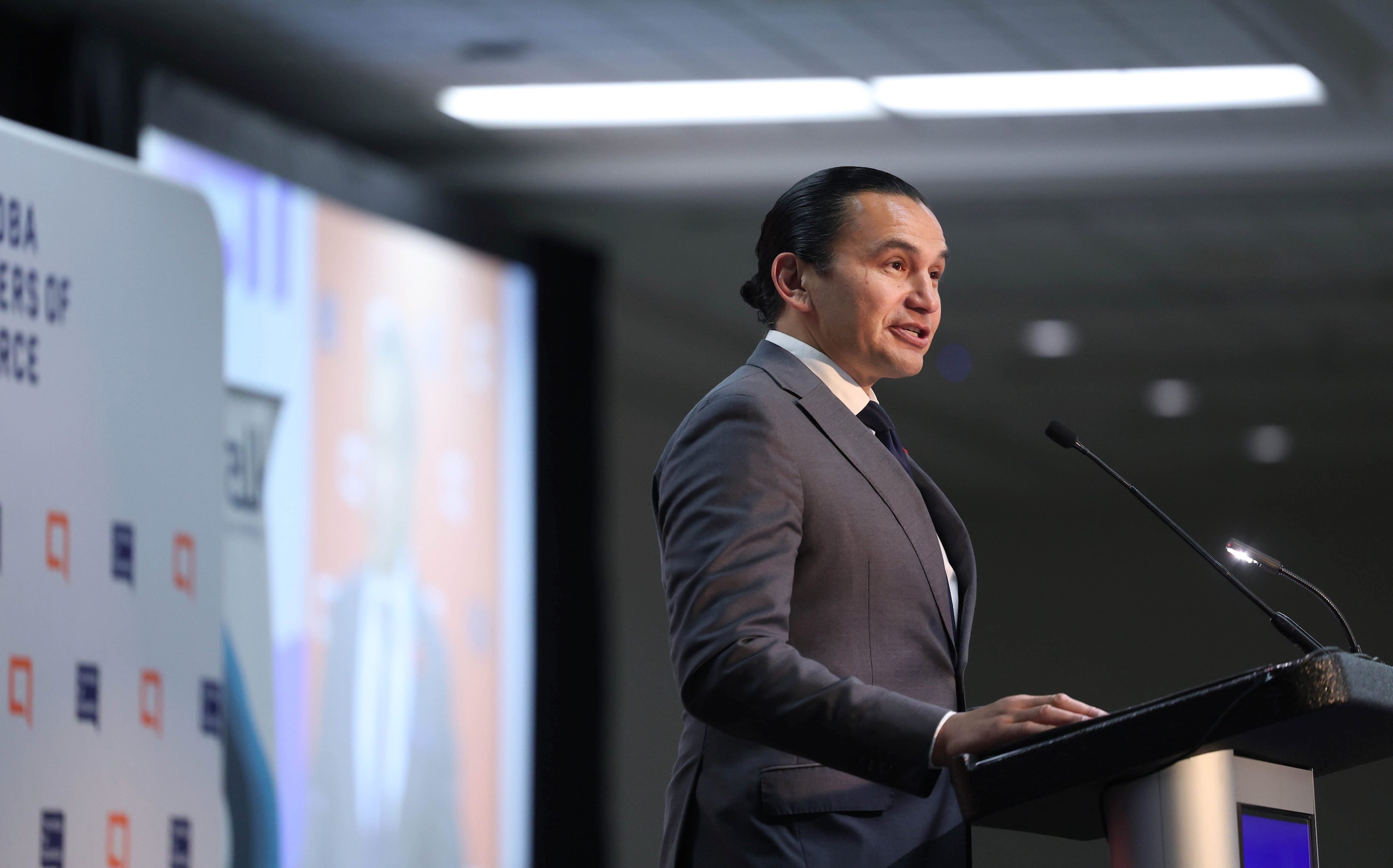 Manitoba Premier Wab Kinew, wearing a dark suit, stands at a podium to speak to the provincial Chambers of Commerce about a new innovation task force report