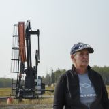 A woman looks off into the distance with an oil and gas rig in a field behind her.