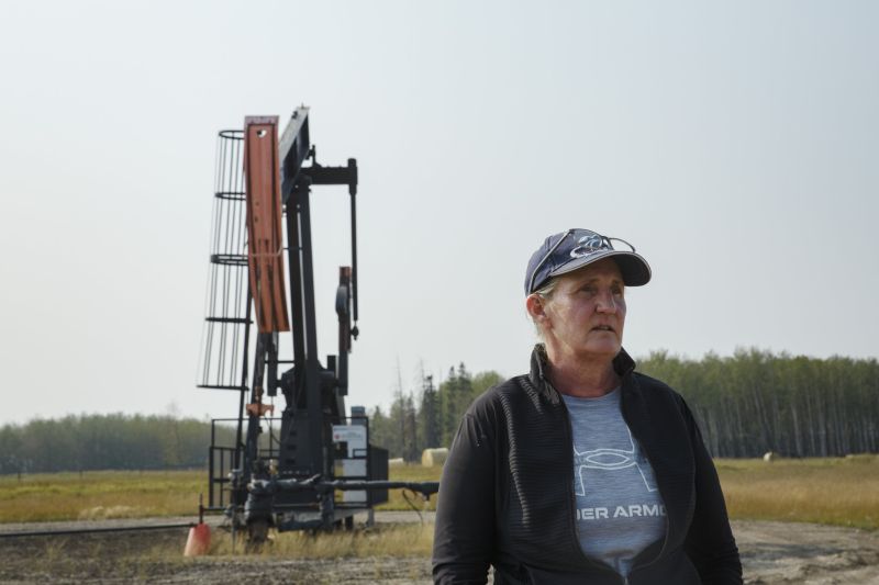 A woman looks off into the distance with an oil and gas rig in a field behind her.