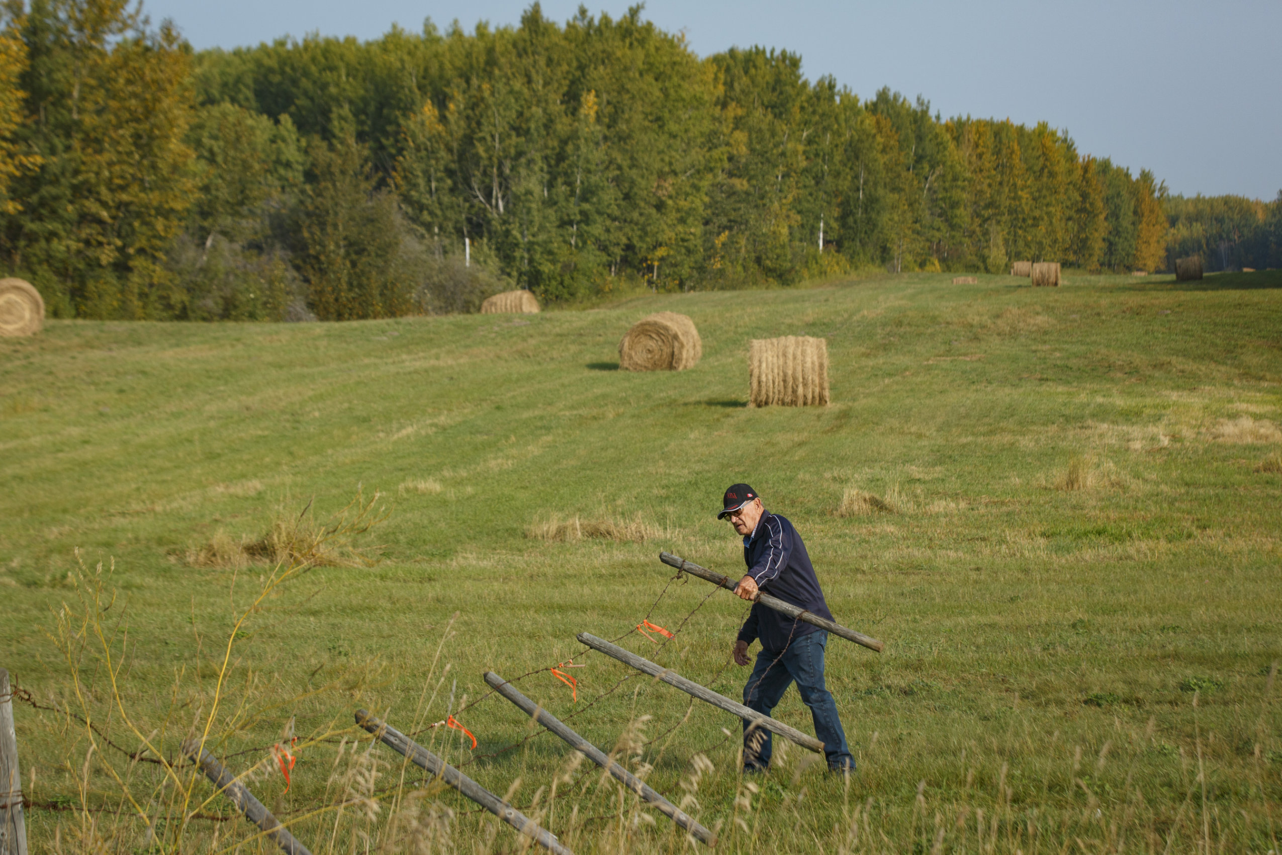 A man pulls a wire fence towards himself in a wide open grassy farm field with hay bales in the distance.