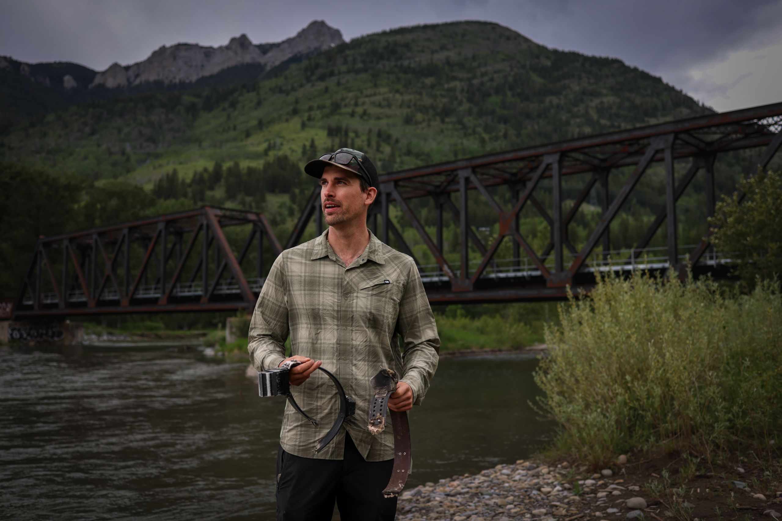a photo of wildlife scientist Clayton Lamb holding damaged GPS collars on the edge of the Elk River near a rail bridge with mountains behind