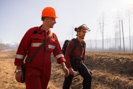 Two men wearing fire protective gear and hard hats walk through a remote area during a prescribed burn.