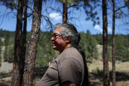 Joe Gilchrist, a Secwépemc firekeeper, stands in profile against a pine forest behind him.