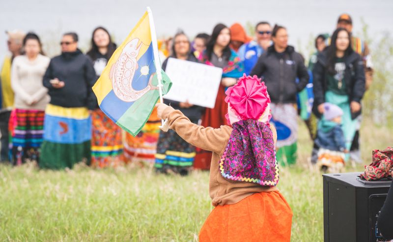 A young girls waves the flag of Kitchenuhmaykoosib Inninuwug First Nation in front of a crowd of mostly women in ribbon skirts