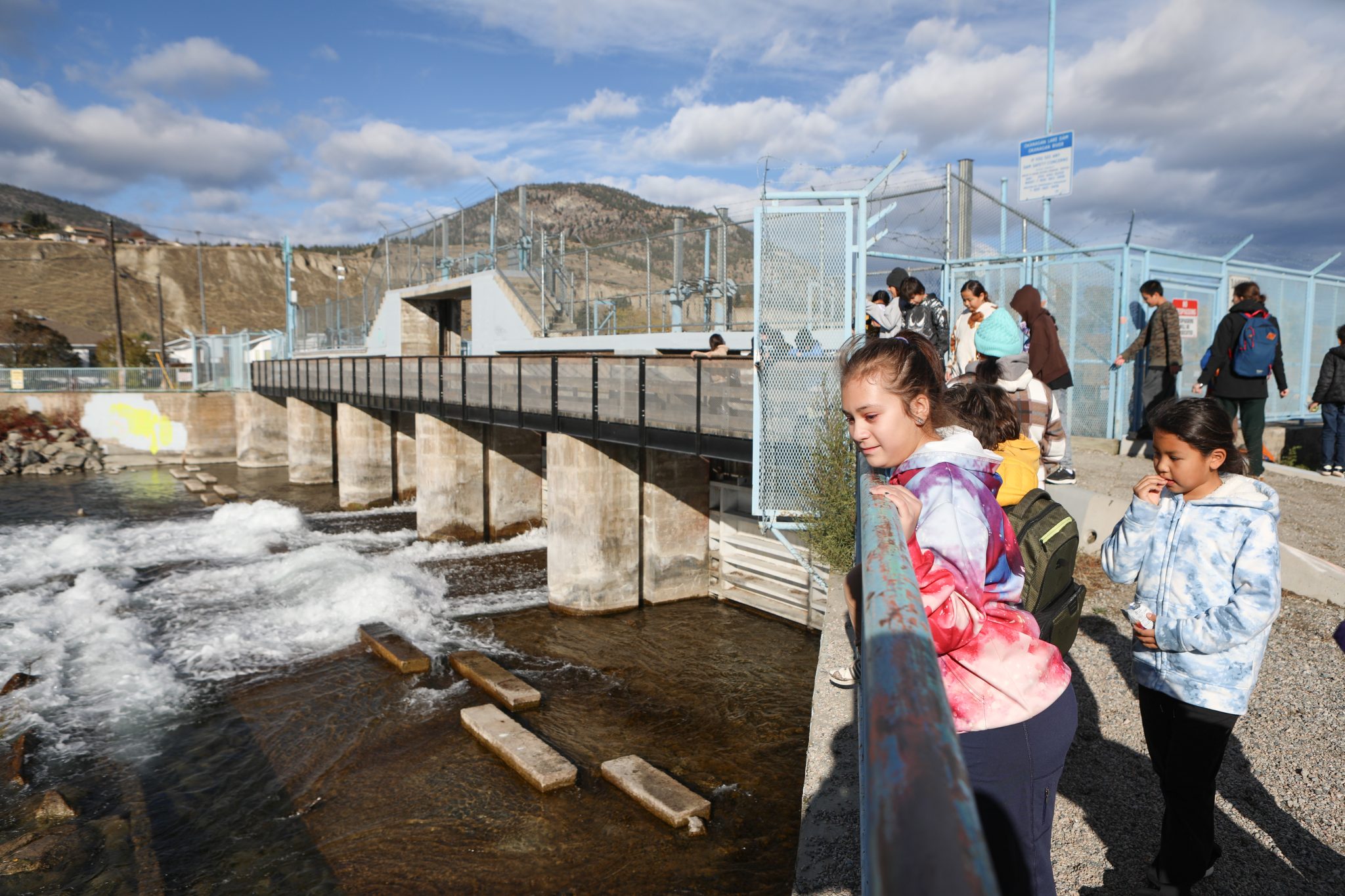 Children lean over a railing and watch as water rushes by after flowing through a dam.