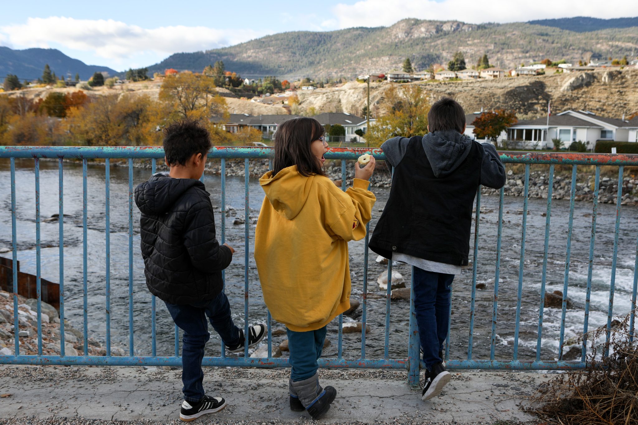 Three children, photographed from behind, look over a blue fence at a set of shallow rapids below a dam.