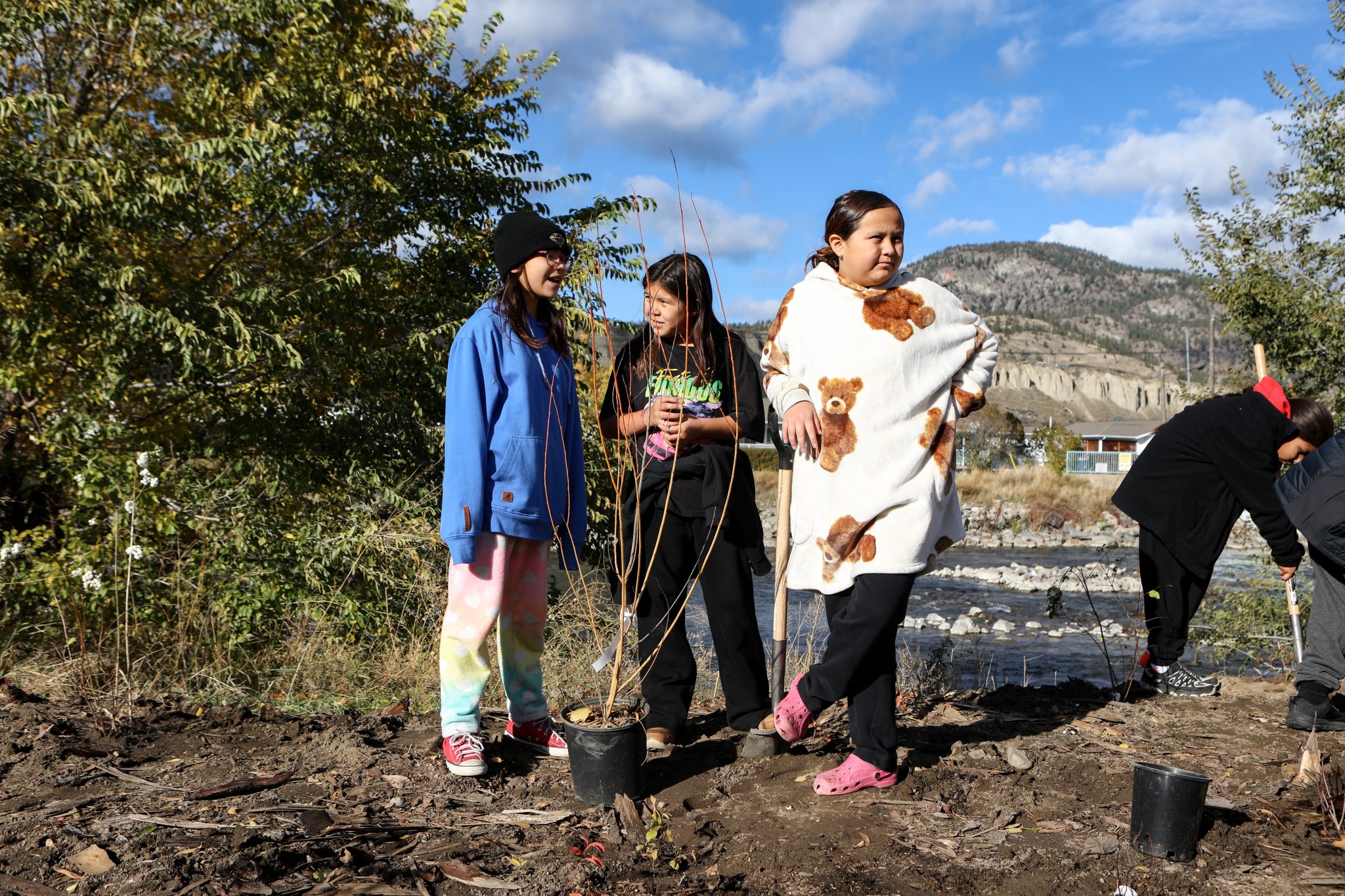 Three children, one leaning on a shovel, stand in front of a potted sapling ready to be planted.