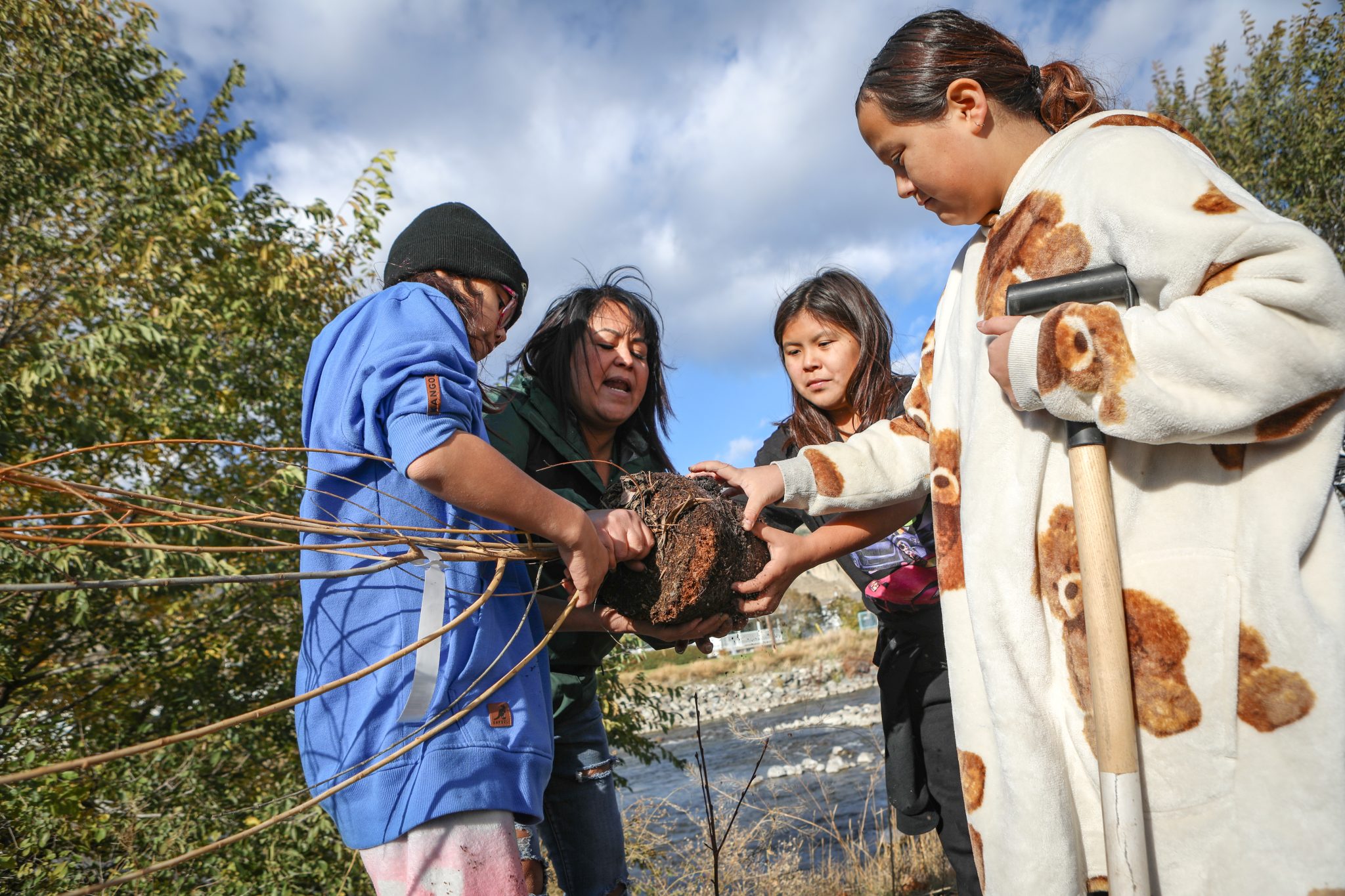 A woman and three children, one of whom is holding a shovel, grab a sapling by its roots.
