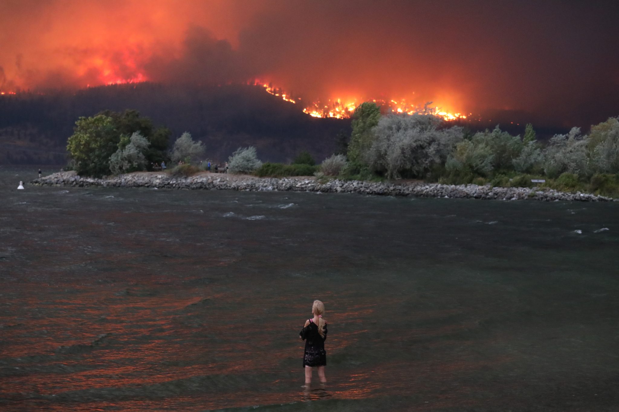 A woman standing in a lake with fire on the hillside in front of her.
