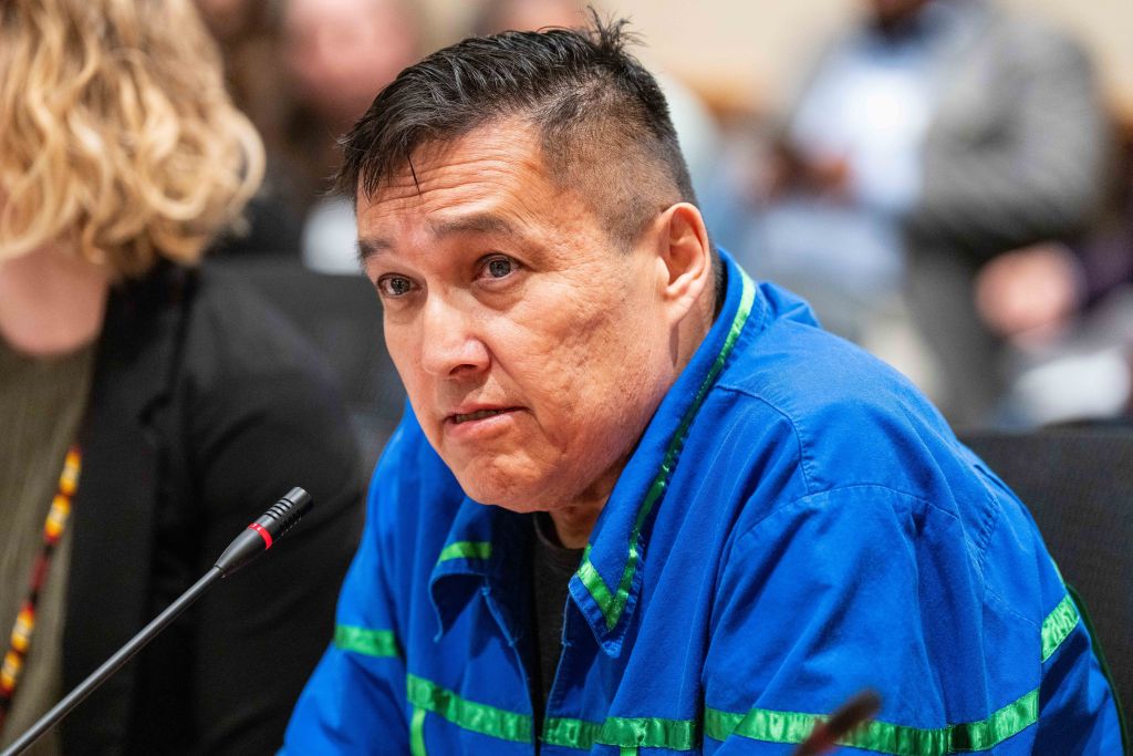 Man in a blue jacket speaks at a microphone at a table.