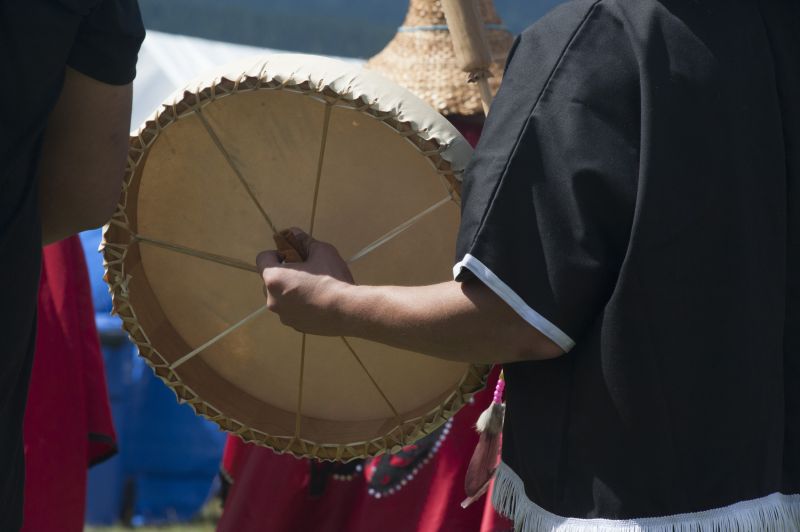 Hand holding moose-hide drum, person wearing a cedar woven hat in background