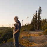 Don Pettit stands at the edge of the ridge on Bear Mountain, facing outward. Behind him a line of wind turbines stretches into the distance, interspersed with trees and other vegetation. There's a sunset glow across the scene