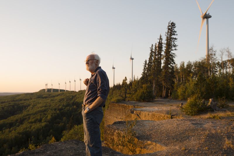 Don Pettit stands at the edge of the ridge on Bear Mountain, facing outward. Behind him a line of wind turbines stretches into the distance, interspersed with trees and other vegetation. There's a sunset glow across the scene