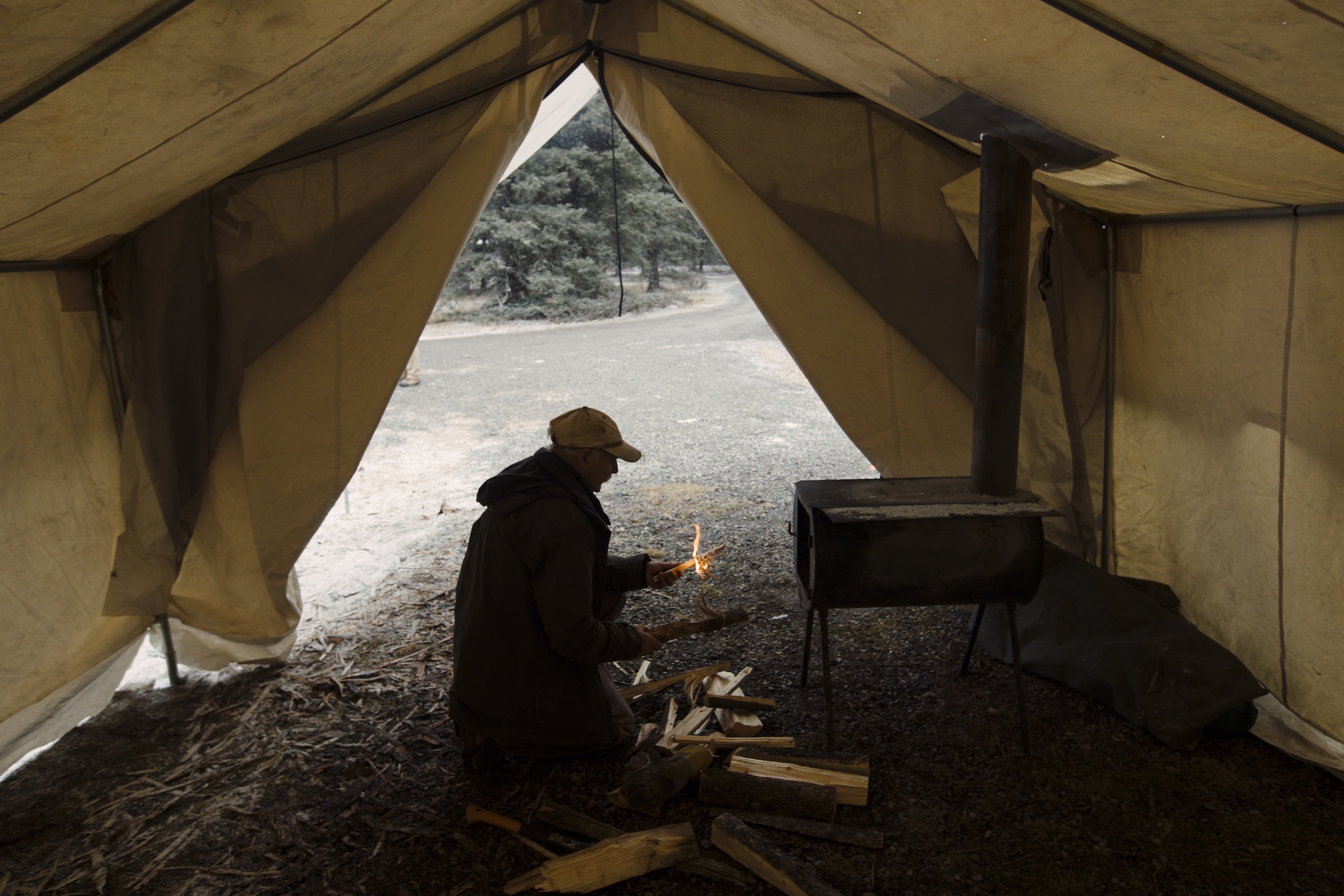A man wearing a baseball cap bends over a small wood stove inside a large tent.