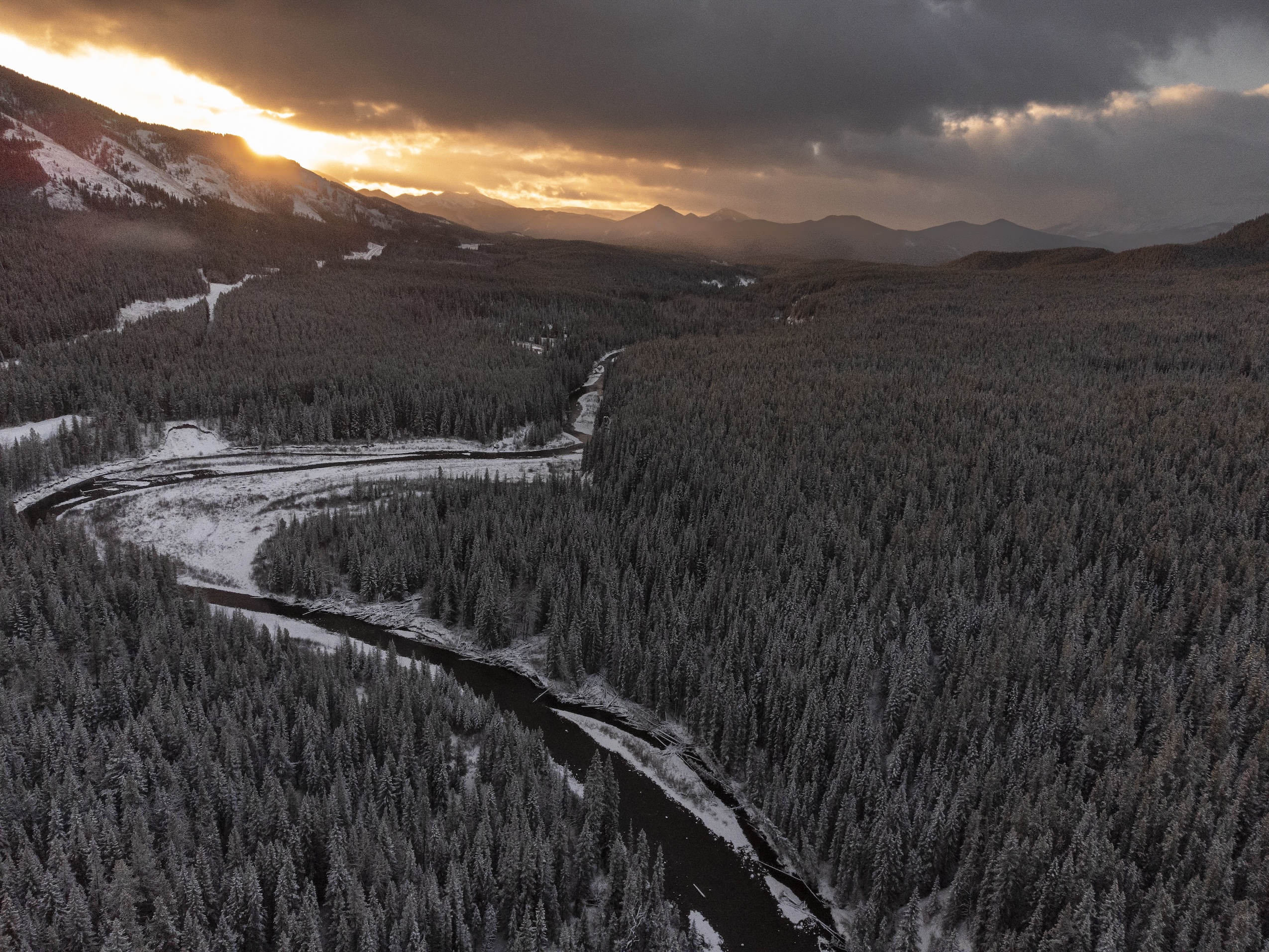A river runs through a forest dusted with snow cover and the sun rising over mountains in the distance.