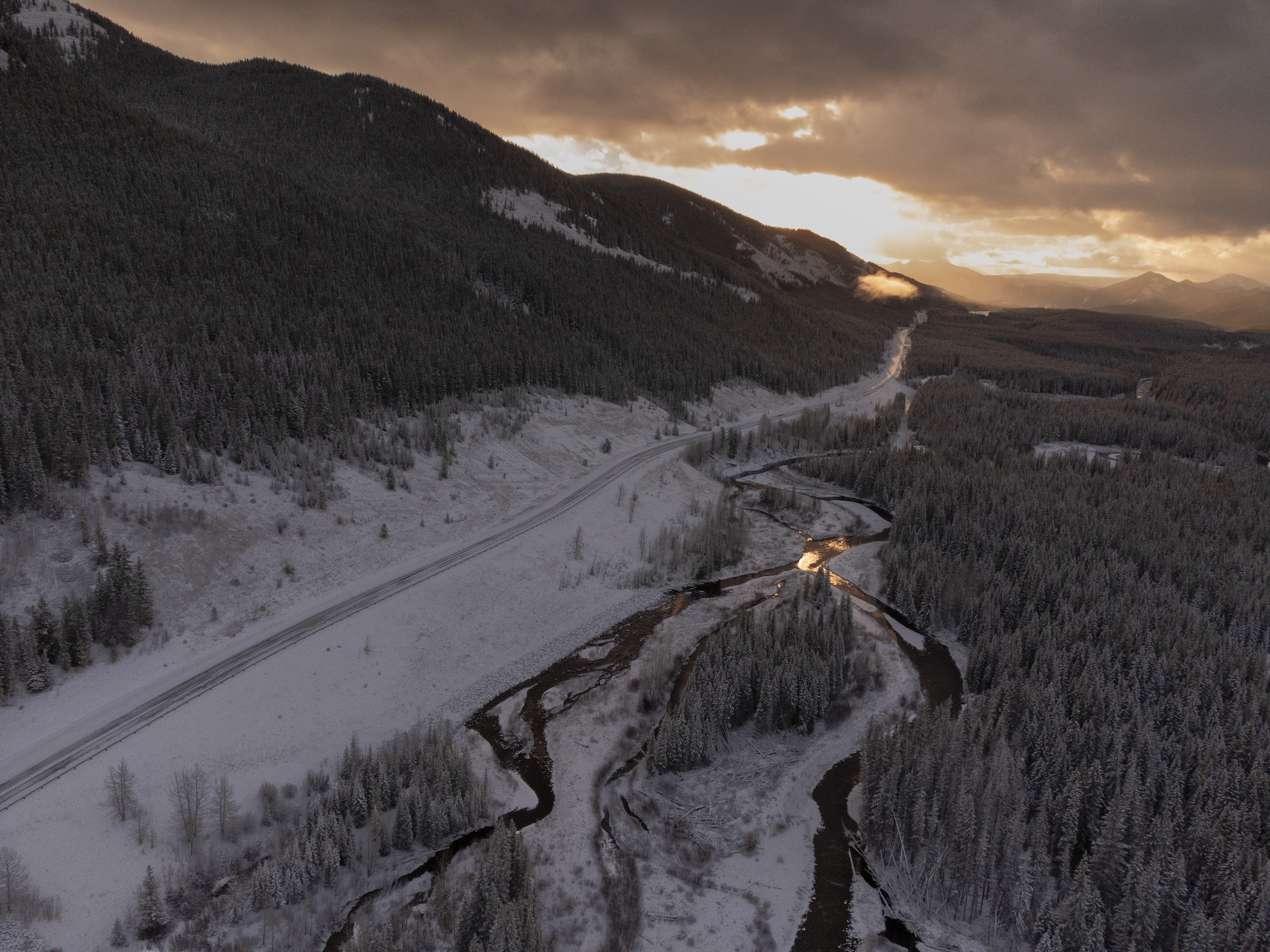 Aerial view of a snow-covered mountain landscape with a highway cutting through it and the sun rising in the distance.