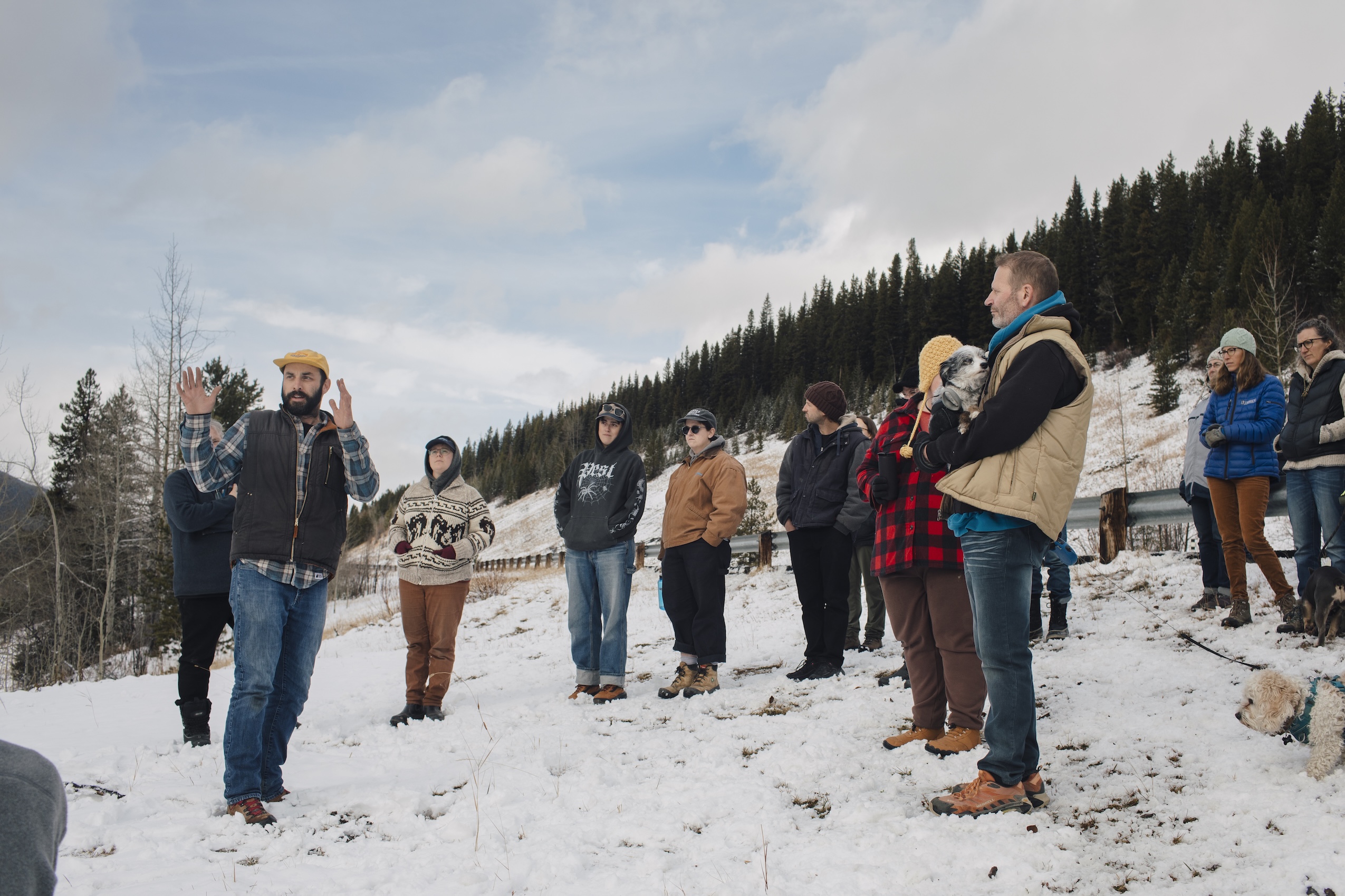A group of people gathered on a snow-covered mountainside along a highway listen to a man speak.