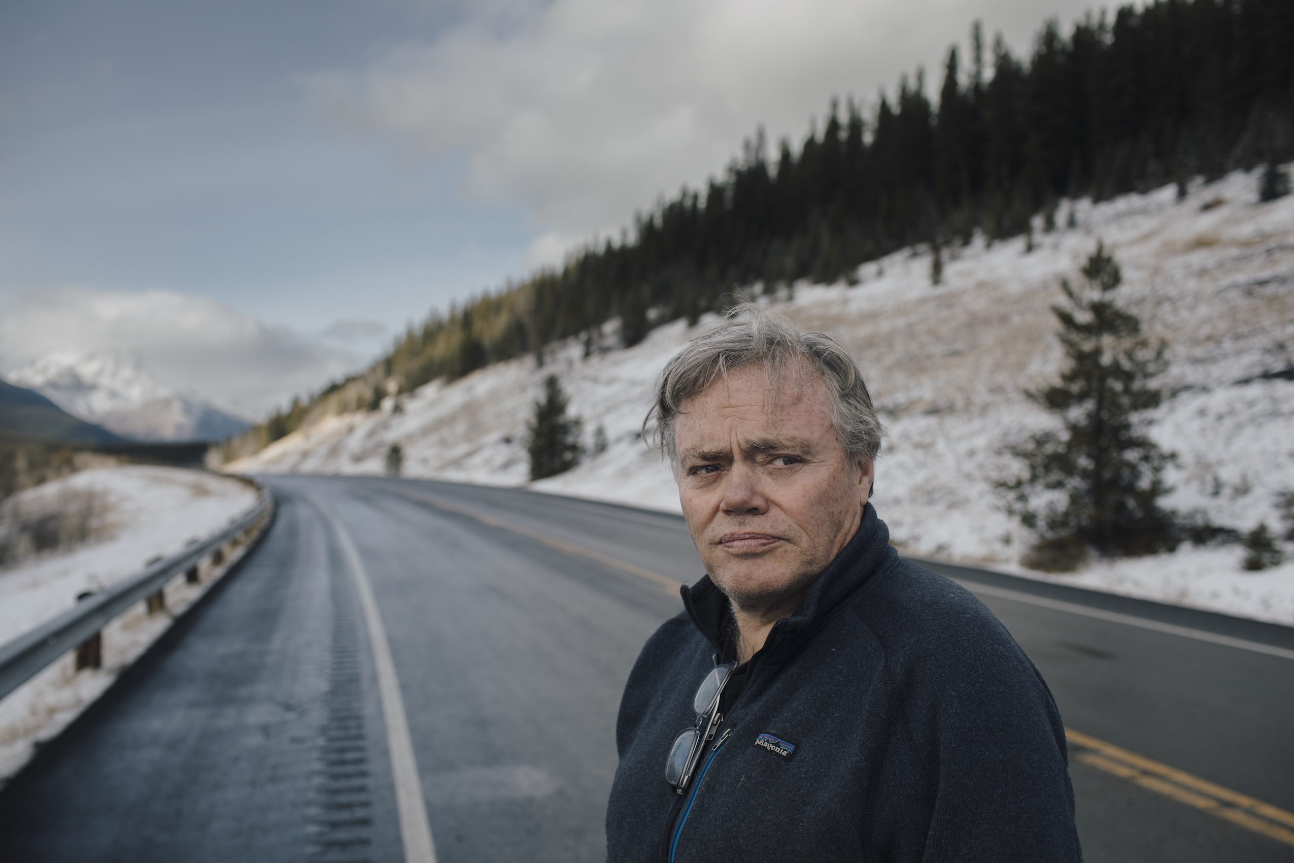A white man with grey hair stands along the side of a highway running through a snowy mountainside.