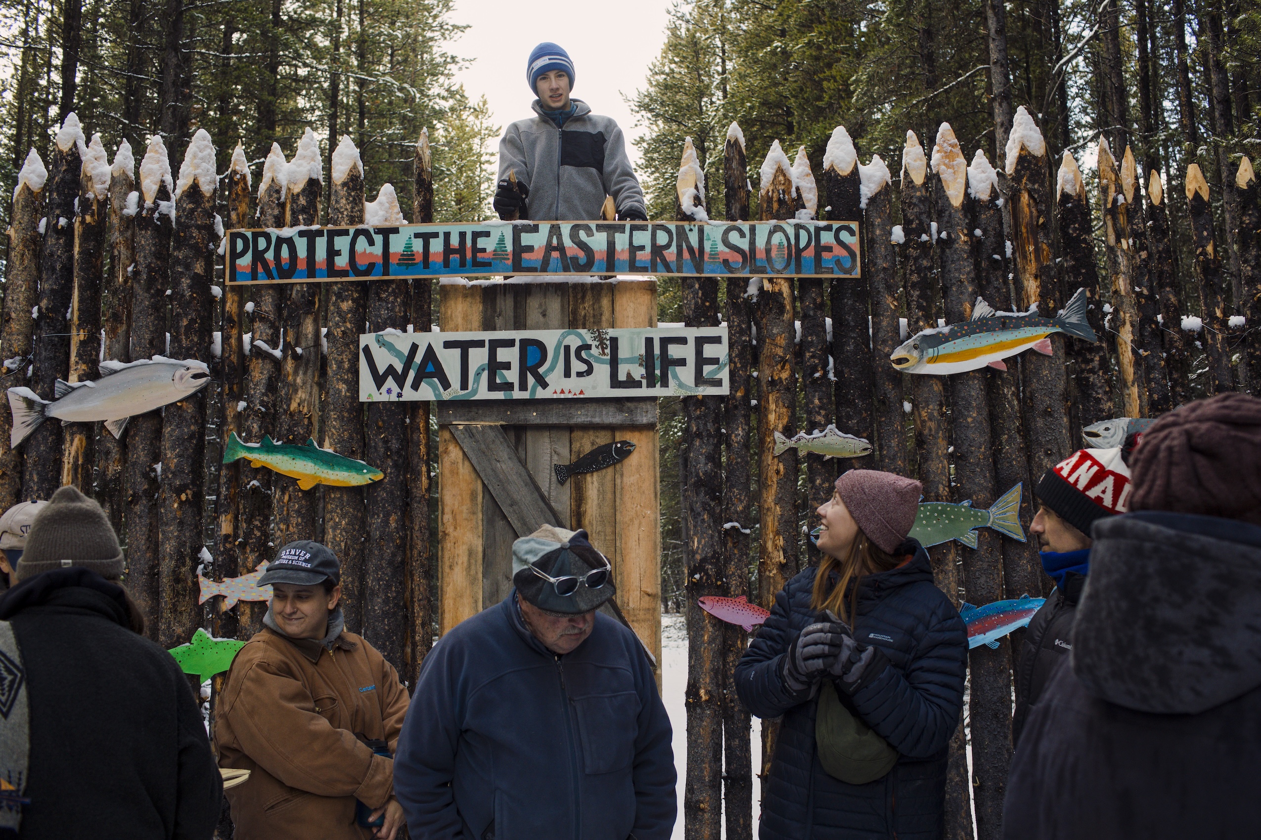 A group of people gathered in front of a wooden gate with a sign reading "Protect the eastern slopes: water is life" on it.