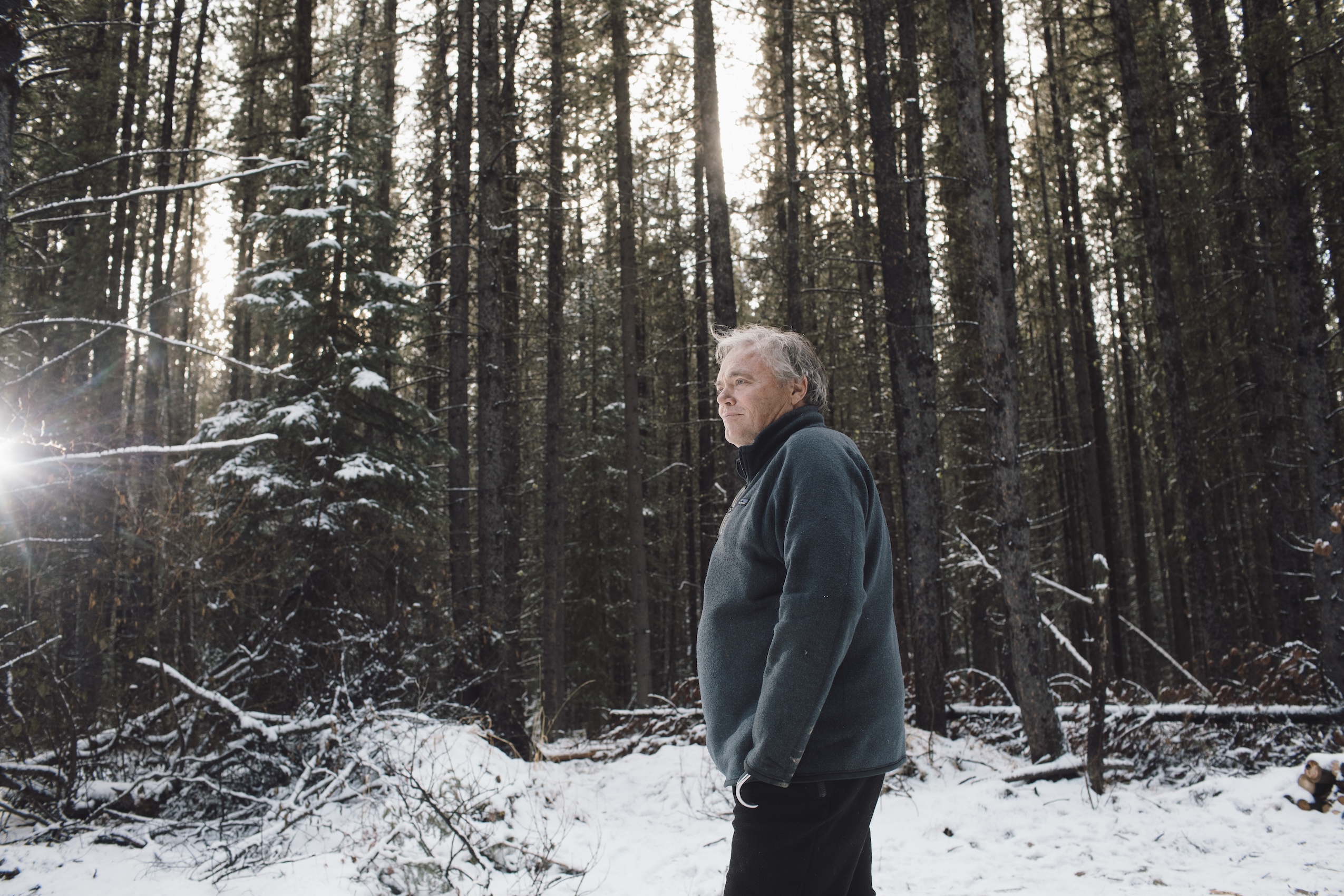 A man with grey hair stands in a snow-covered forest.
