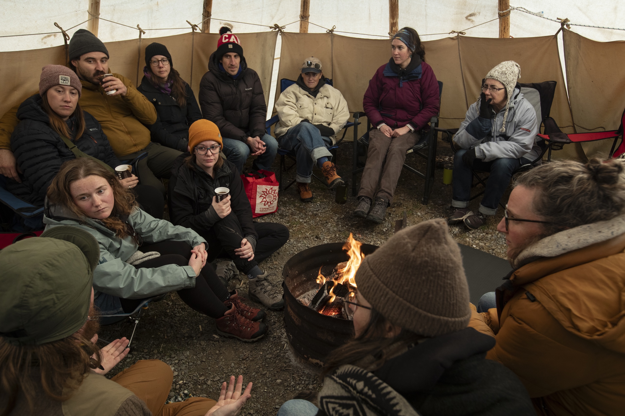 A group of people dressed in warm winter clothing gather around a fire pit inside a tent.