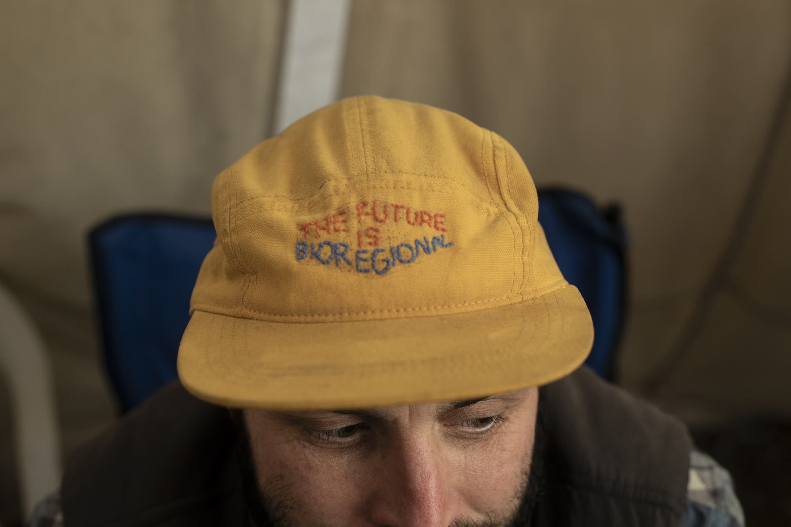 Close-up of a man's yellow baseball hat with the words "The future is bioregional" stitched across it.