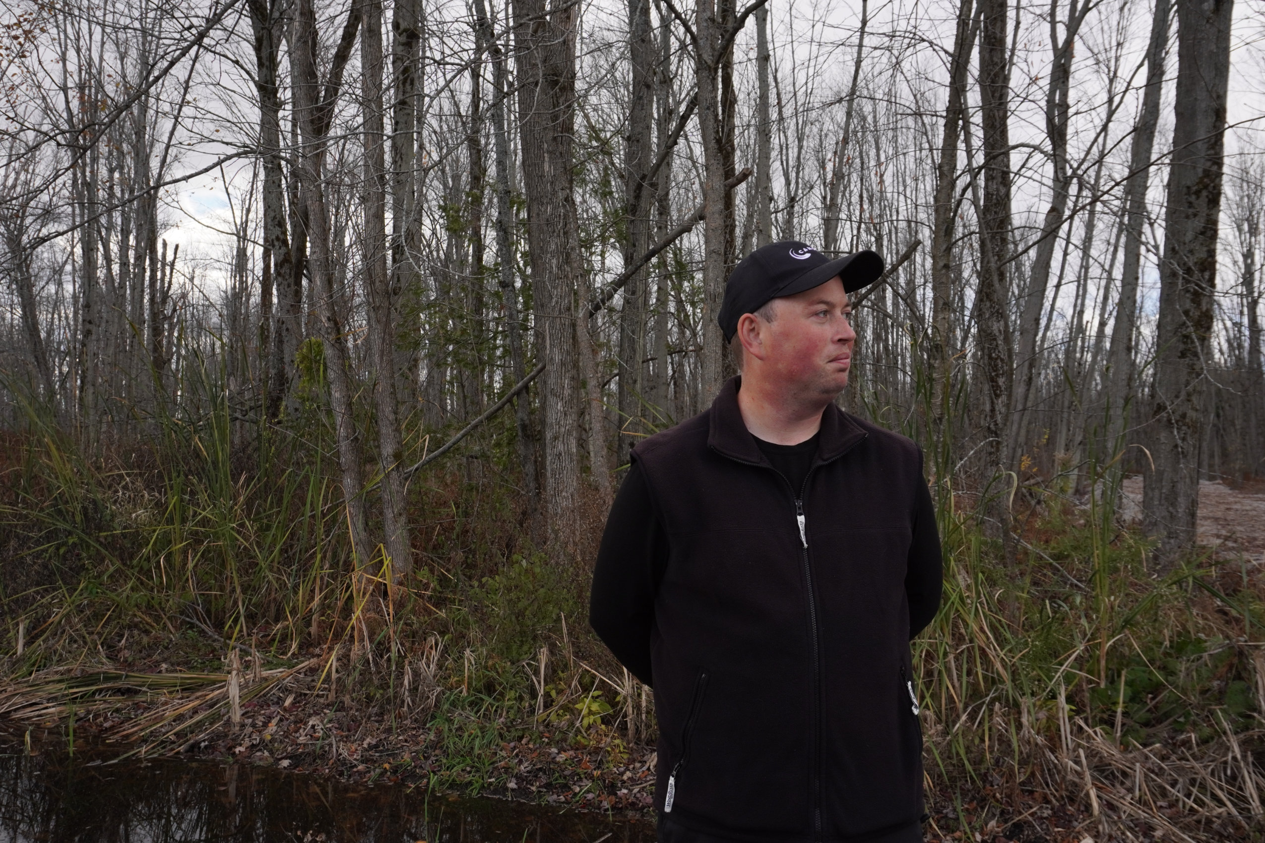 Matt Thomson, wearing a black fleece vest and black baseball cap, looks toward the right, with a treed wetland in the background.