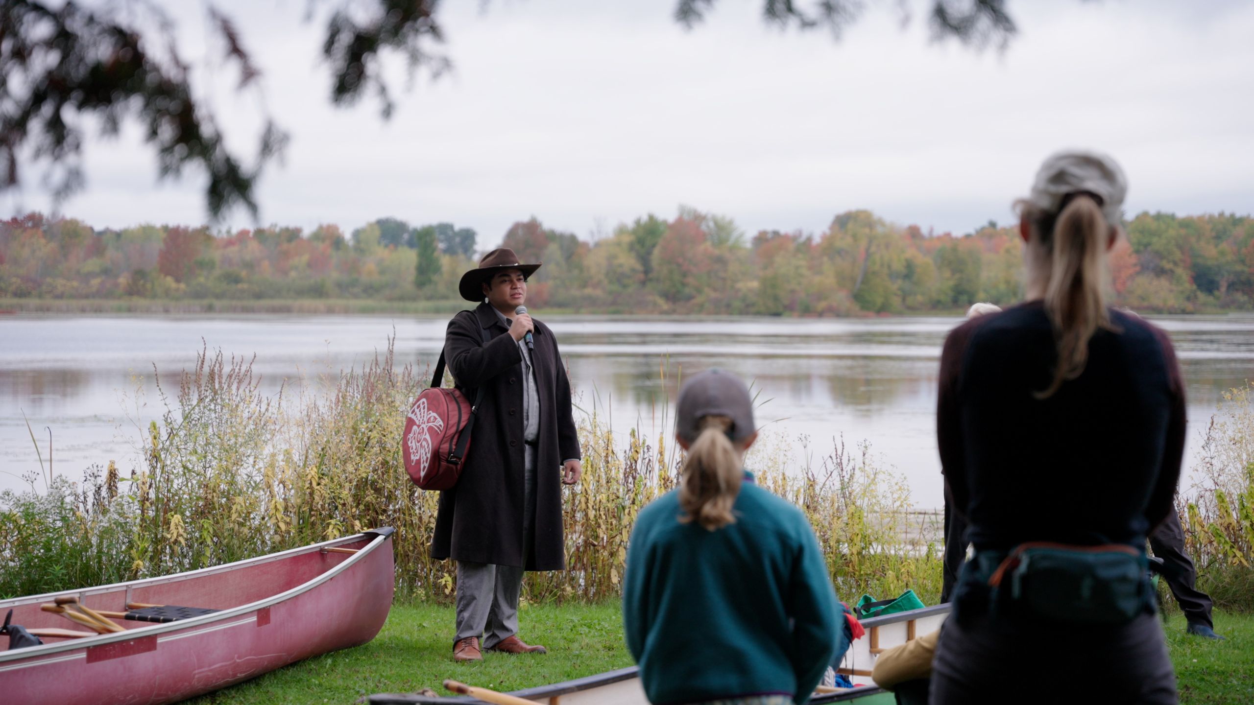 Man outside speaks to listeners next to a canoe in front of a lake.