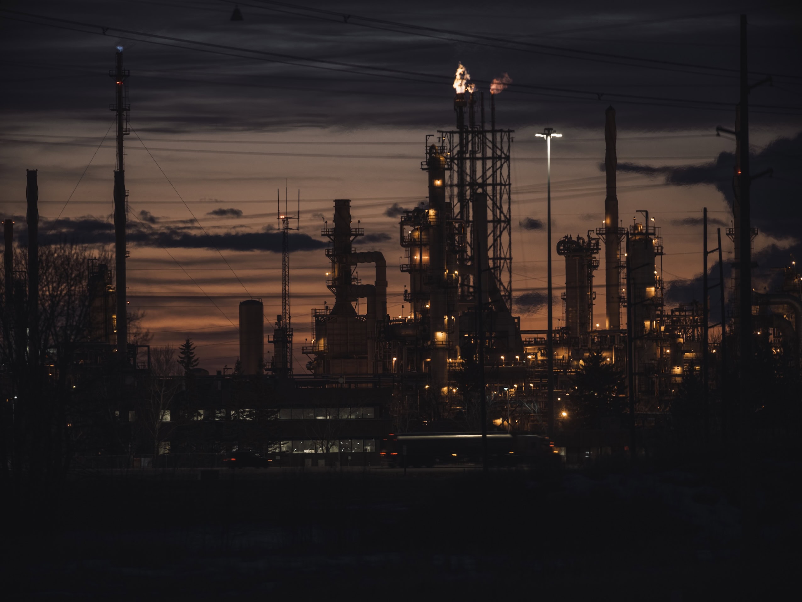 An oil and gas refinery is silhouetted against a dark orange sky.