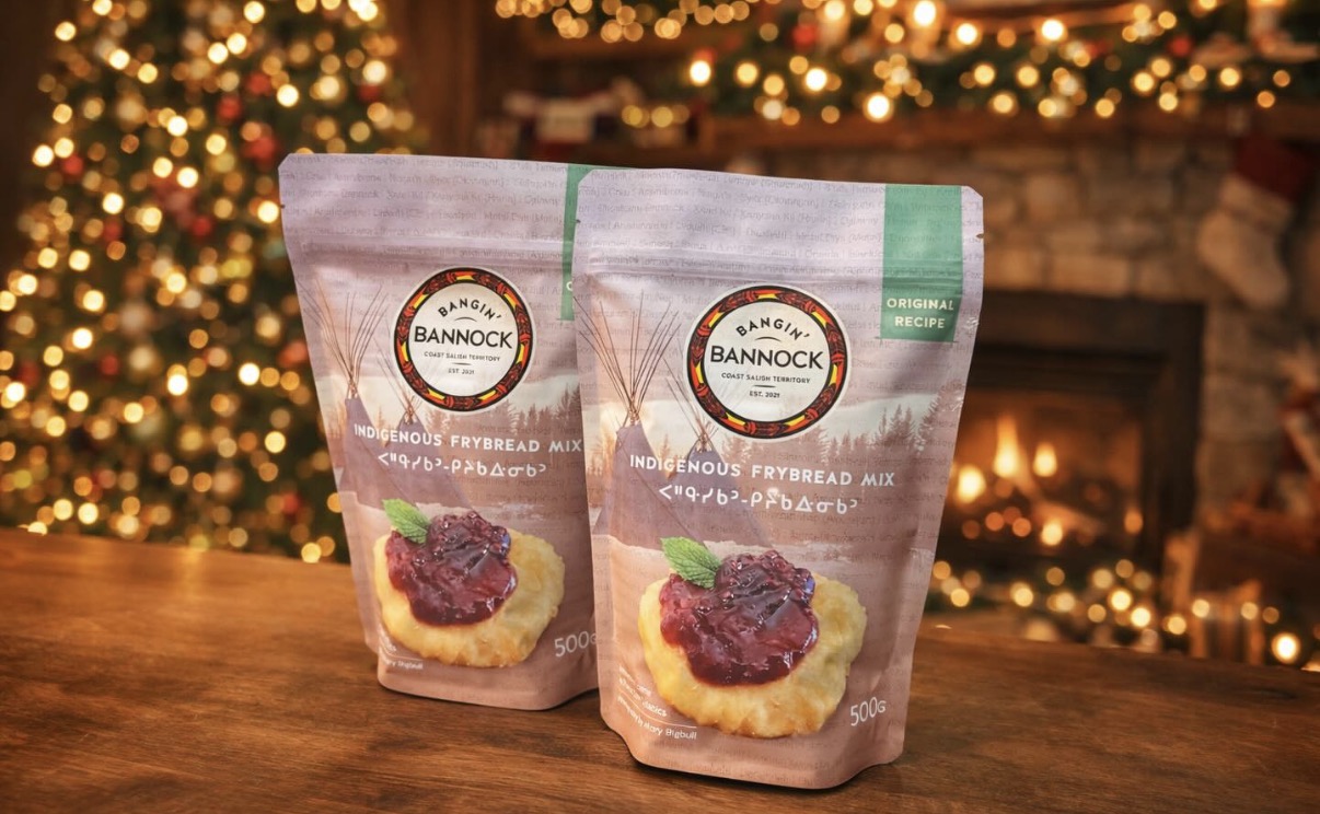 Two bags of Bangin' Bannock mix sit in front of a Christmas tree, Christmas lights and a fireplace. The bags are pink, with a picture of berries on bannock, and have Cree language on the front.