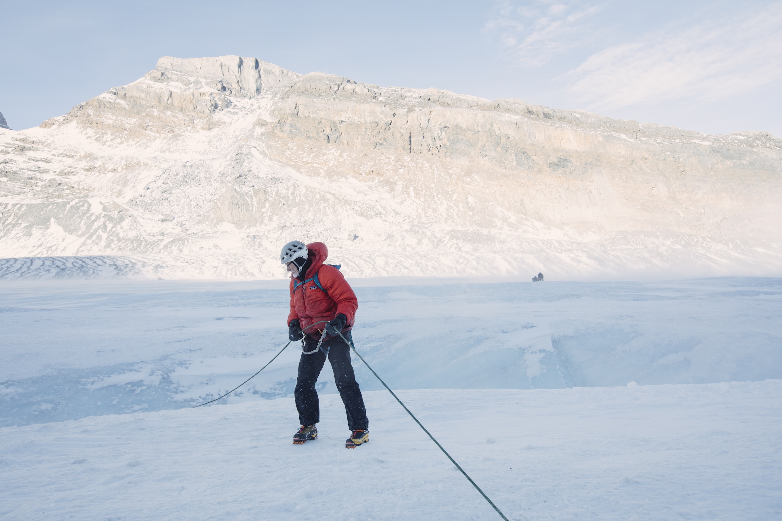 A man with a grey beard wearing a helmet and red jacket is attached to ropes beginning his decent into a glacier moulin
