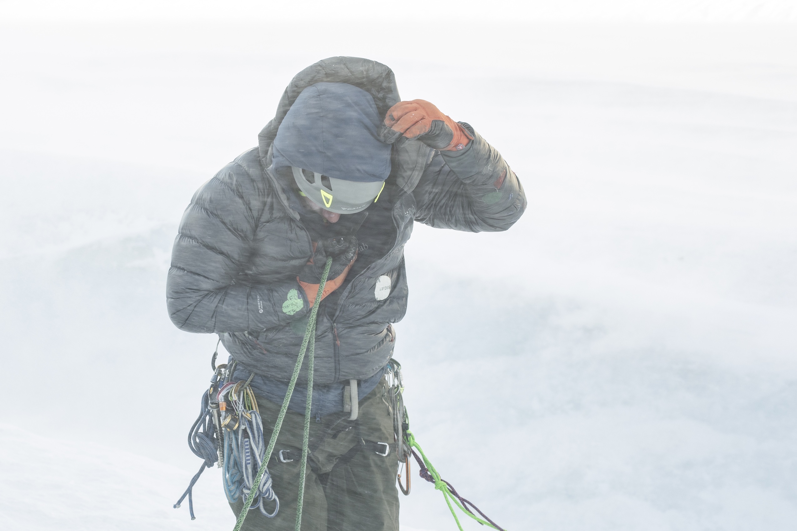 A person wearing a black jacket and helmet and climbing gear covers his head and speaks into a walkie talkie with snow in the background