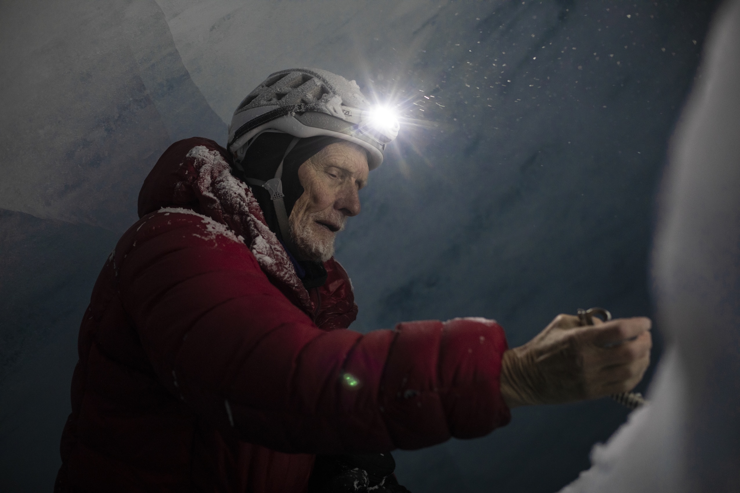 A man with a grey beard wearing a helmet and headlamp that's turned on and a red jacket stands on the inside of a glacier.