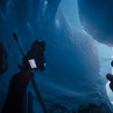 Two people stand with a tripod wearing helmets and headlamps inside of a glacier