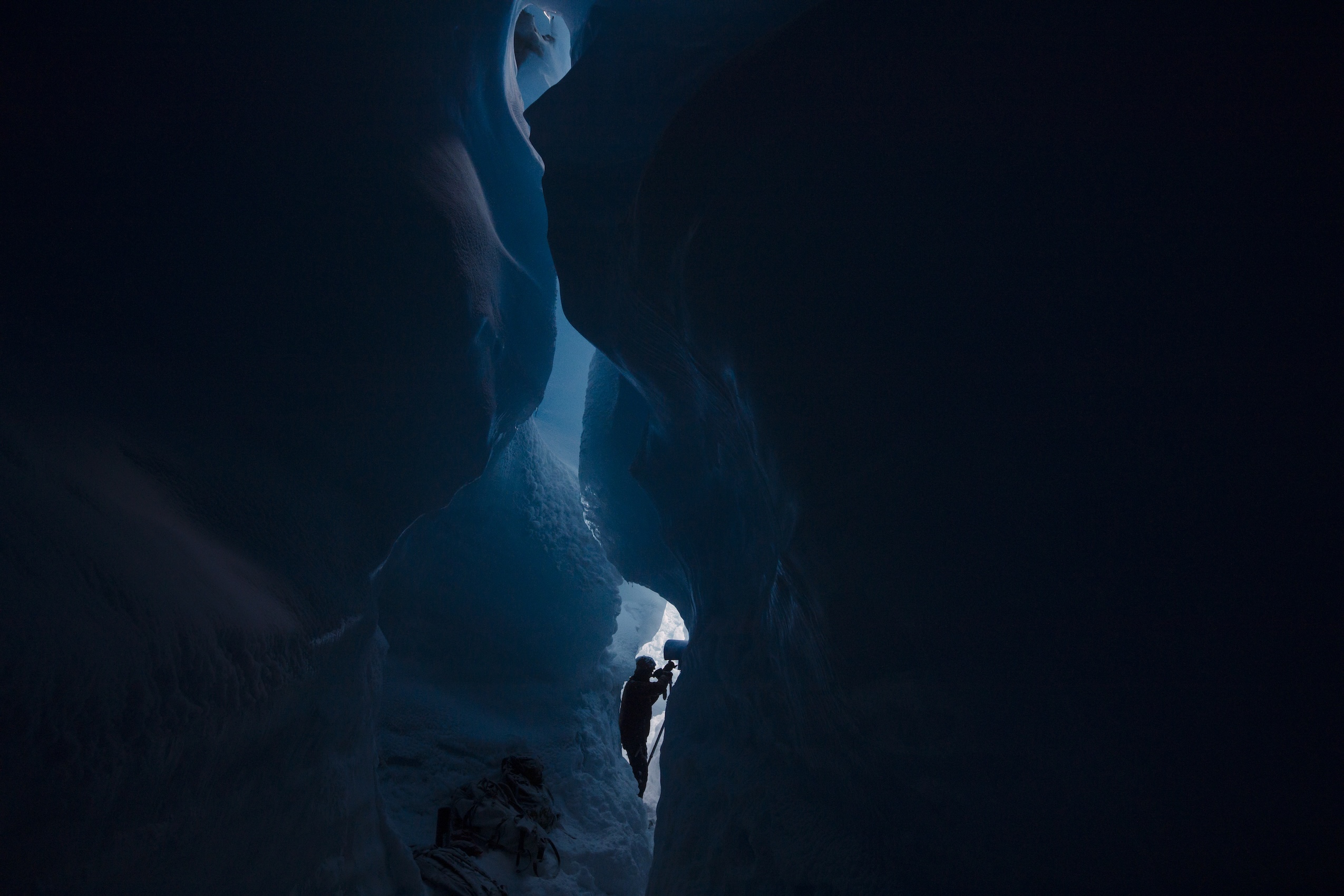 The curvy and icy walls of a glacier are in the foreground with a person holding a camera seen deep in the crack