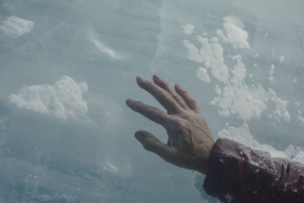 The right hand of a person with wrinkled skin touches a slab of glacial ice