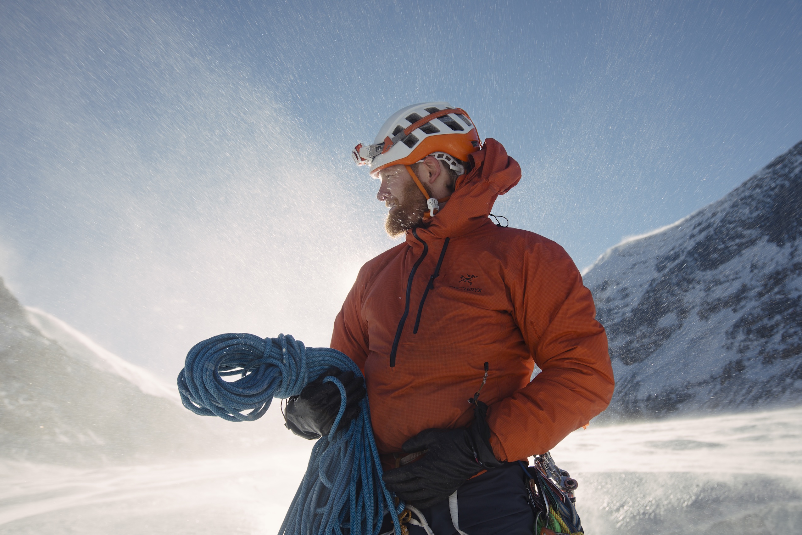 A man with a red beard wearing a white helmet with a headlamp and a red jacket holds a blue rope in front of a glacier