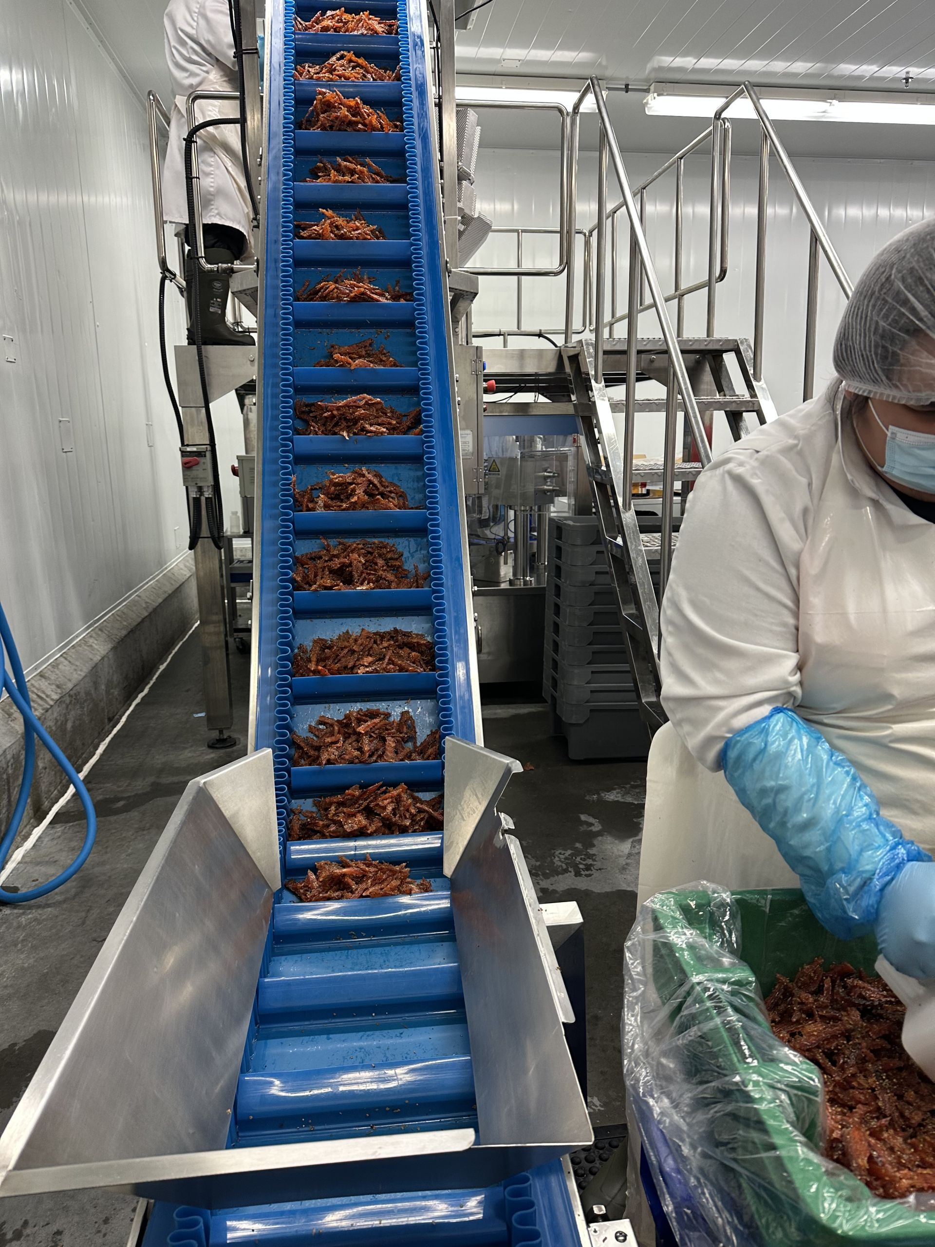 An Authentic Indigenous Seafood jerky machine has a long line of fish coming down a conveyer belt. On the left, a worker wears a hair net, face mask and gloves and processes fish.