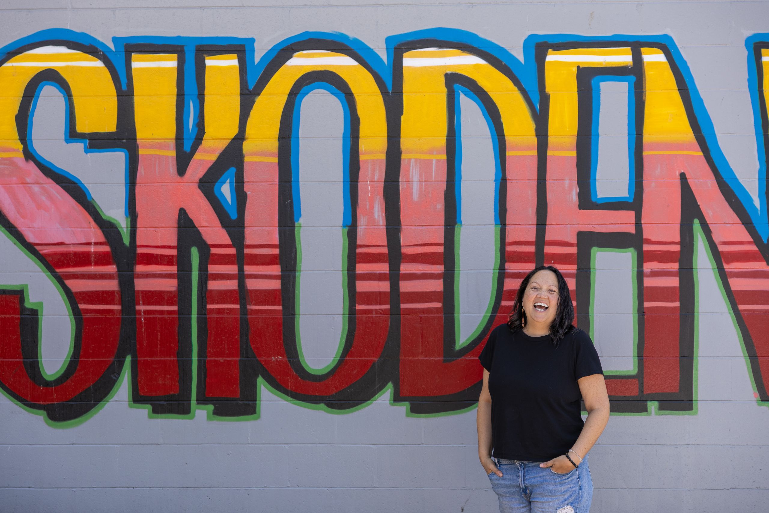 Jordan Hocking laughs, looking off camera, standing in front of a large piece of wall art that says SKODEN in all caps, painted in an orange-to-red gradient.