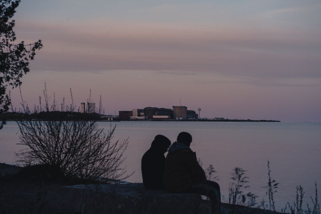 The Pickering Nuclear plant is in the horizon of a sunset sky as two people sit on the Lake Ontario shores