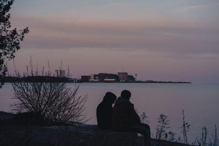 The Pickering Nuclear plant is in the horizon of a sunset sky as two people sit on the Lake Ontario shores