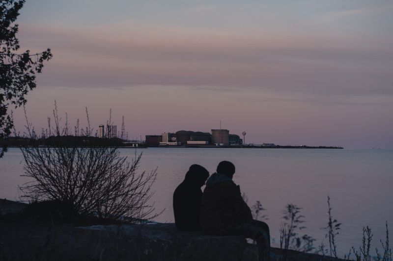 The Pickering Nuclear plant is in the horizon of a sunset sky as two people sit on the Lake Ontario shores