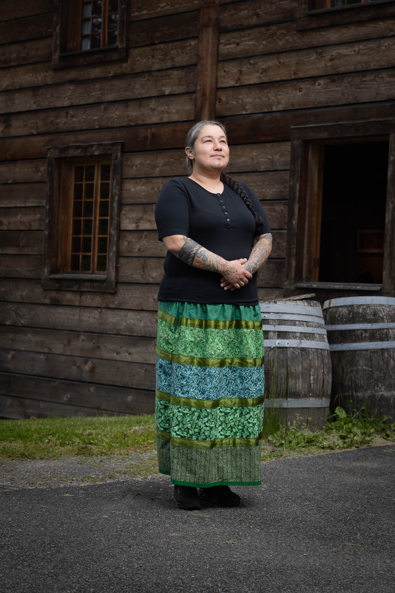 Sarah Meconse Mierau stands in front of a wooden wall, two barrels off to the right. She raises her head towards the sunlight, facing to the right slightly. Her hands as clasped in front of her, and she wears a vibrant green ribbon skirt. She has a peaceful expression.
