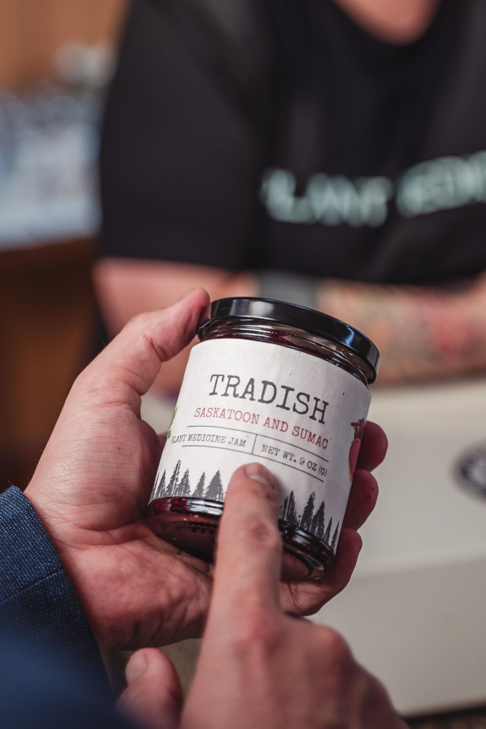A close-up of a jar of Tradish jam in someone's hands. It says Tradish, Saskatoon and Sumac, plant medicine jam, 9 oz.