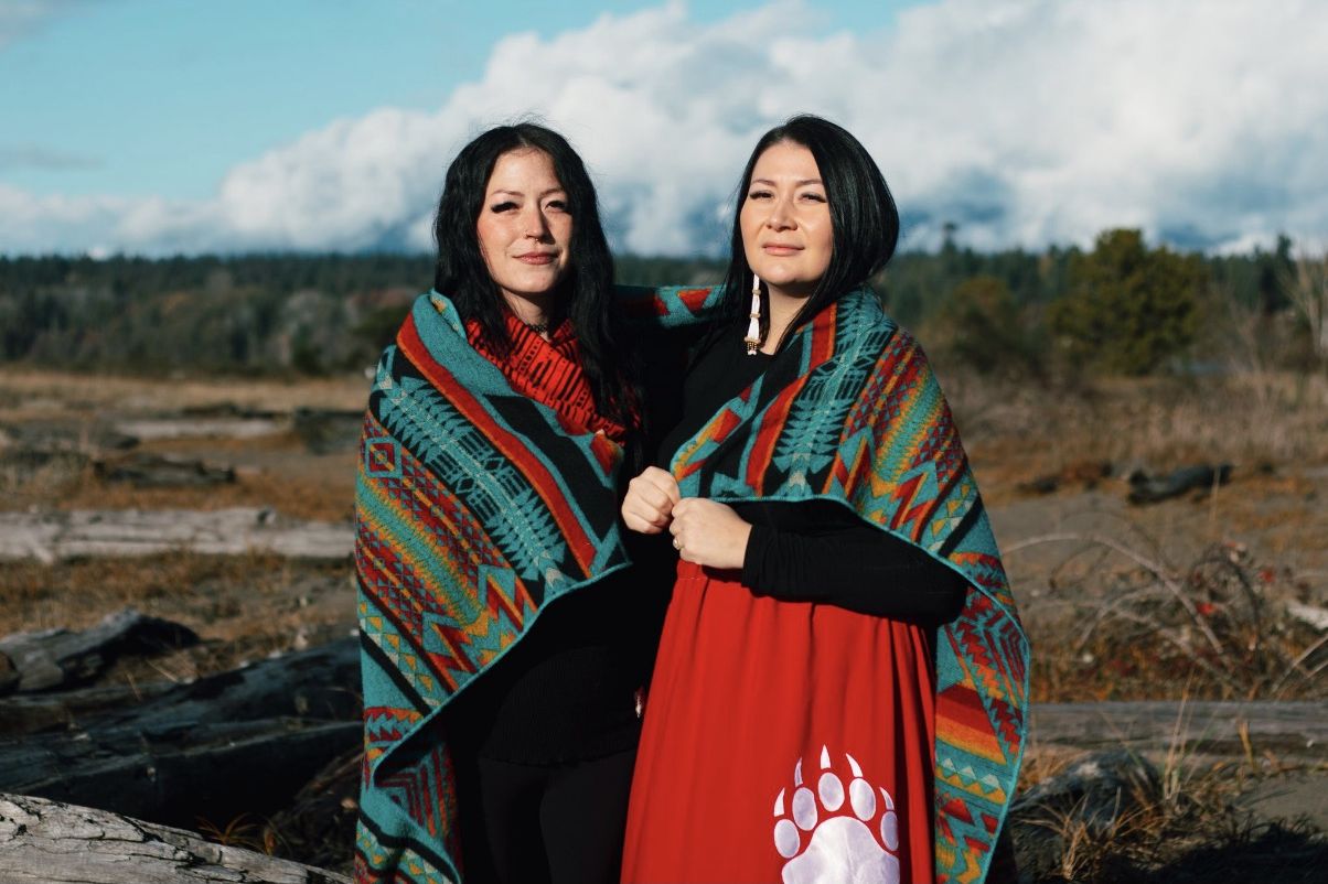 Bangin' Bannock co-founders Kelsey Coutts and Destiny Houshte wear blankets on their shoulders, looking into the camera, a field and trees extending behind them.
