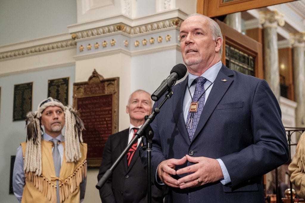 Premier John Horgan standing outside the legislative chamber, flanked by Green Party MLA Adam Olsen in traditional regalia and former Indigenous Relations and Reconciliation Minister Scott Fraser