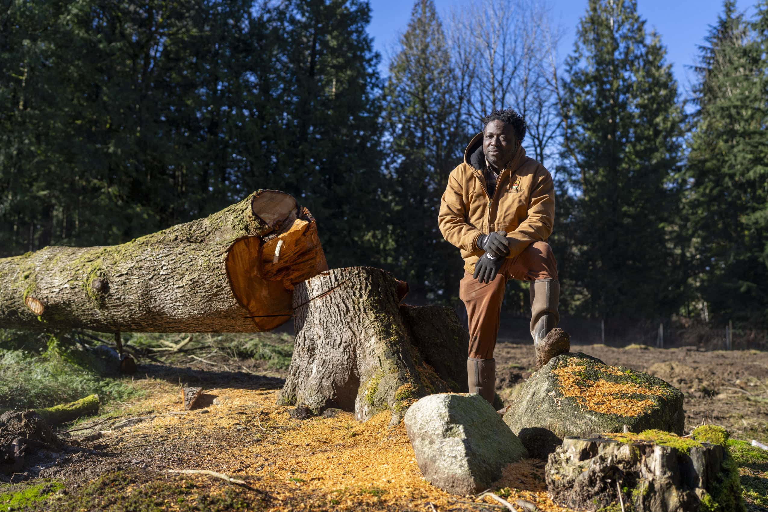 Toyin Kayo-Ajayi stands in the sun, one foot up on a rock and leaning on his knee, facing slightly to the left where the sun is coming in, but looking straight into the camera with a calm expression. Beside him, a tree that has just been cut down rests by the leftover stump.