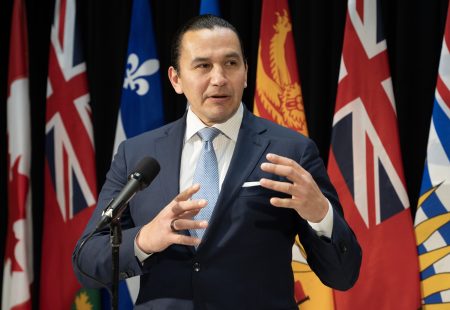 Manitoba Premier Wab Kinew speaks into a microphone in front of a row of provincial flags.