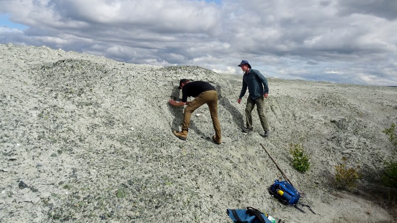 Two men stand on a grey waste rock pile in the Yukon under cloudy skies
